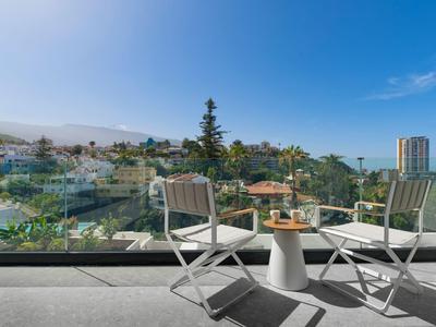 Zwei Stühle und ein Tisch auf einem Balkon mit Blick auf einen Pool und umliegende Gebäude bei klarem Himmel.