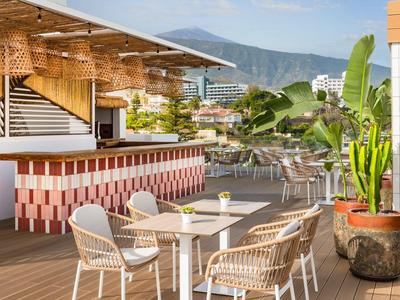 Offener Hotelterrassenbereich mit Tischen, Stühlen und Bar vor Bergkulisse bei sonnigem Wetter.