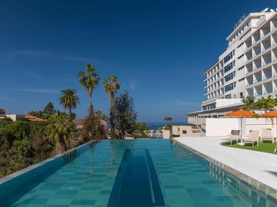Moderner Hotelpool mit Palmen, Liegestühlen und Blick auf das Meer unter klarem Himmel.