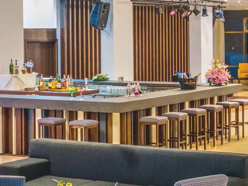 Moderne Bar mit Holzbarhockern, grauer Sitzbank, Sesseln und bunten Cocktails auf Tischen.