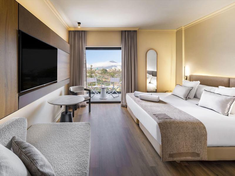 Modernes Hotelzimmer mit großem Doppelbett, Sofa, Flachbildfernseher und Balkon mit Meerblick.