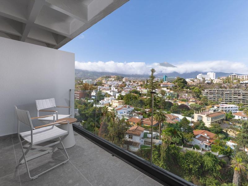 Balkon mit weißen Stühlen und Glasgeländer, Blick auf Stadt mit grünen Bäumen und Bergen im Hintergrund.