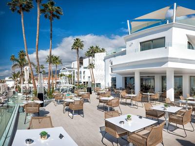 Moderne Terrasse mit vielen Tischen und Stühlen, weißen Gebäuden und Palmen unter blauem Himmel.