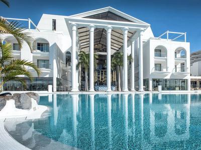 Weißes Hotel mit großen Säulen und blauem Pool im Vordergrund unter klarem Himmel.