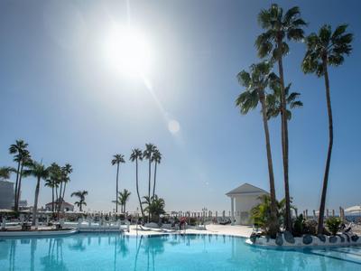 Schwimmbecken mit Palmen, Sonnenschirmen und hellem Himmel bei starker Sonneneinstrahlung.