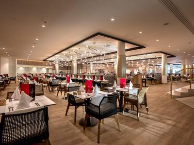 Großer moderner Speisesaal mit Holzboden, Tischen mit weißen Tüchern und schwarzen Stühlen, warmes Licht.