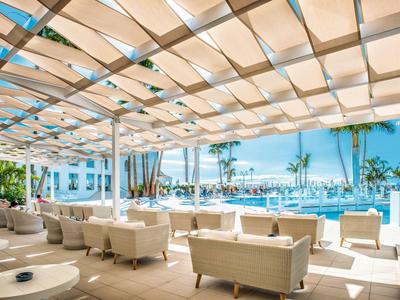 Otwarta przestrzeń hotelowa z białymi meblami, basenem i widokiem na morze w słoneczny dzień.