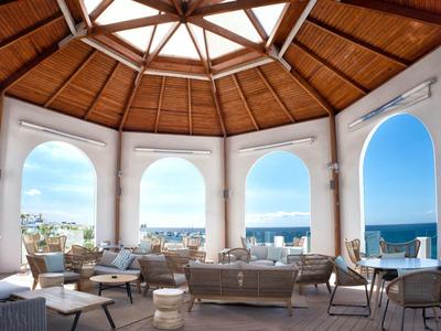 Offener Raum mit Holzdach, großen Bögen und Blick auf das Meer mit Sitzgruppen und Tischen.