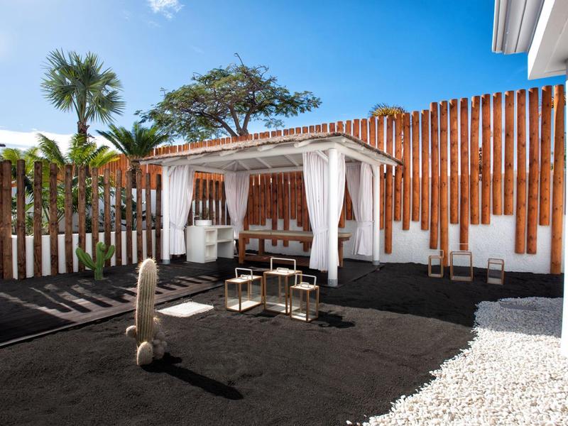 Moderner Garten mit Holzsichtschutz, schwarzem Bodenbelag, weißer Pergola und Sitzgruppe unter blauem Himmel.