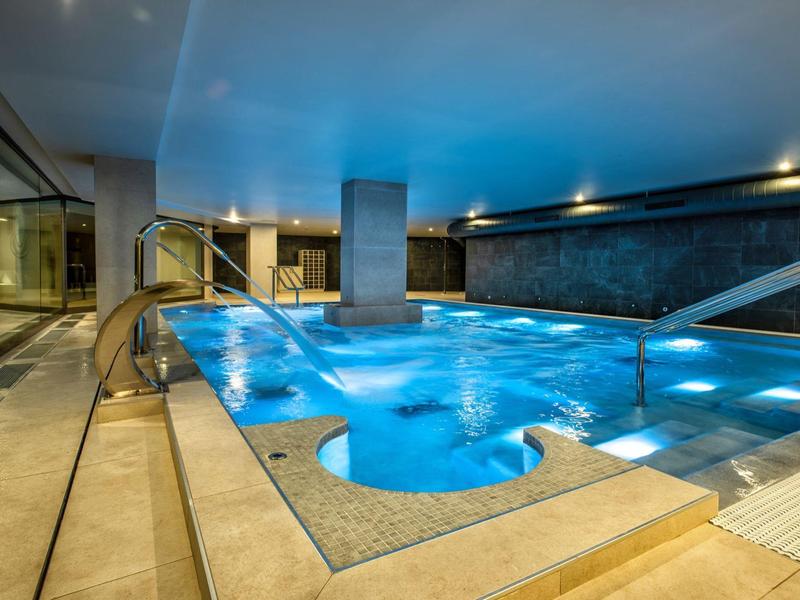 Moderner Innenpool mit blauer Beleuchtung, weißem Deck und glasierten Wänden bei Dämmerung.