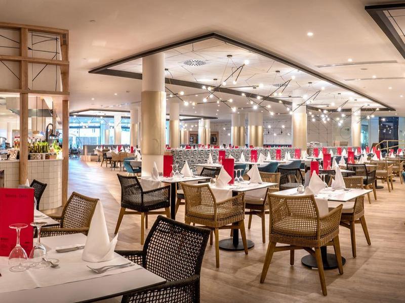 Großes Restaurant mit weißen Tischtüchern, roten Servietten und hellem Holzmobiliar in warmem Licht.