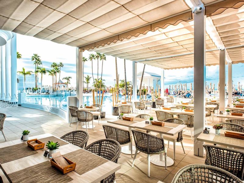 Sonnige Terrasse mit Tischen, Stühlen und Blick auf Palmen und Pool im Hintergrund.