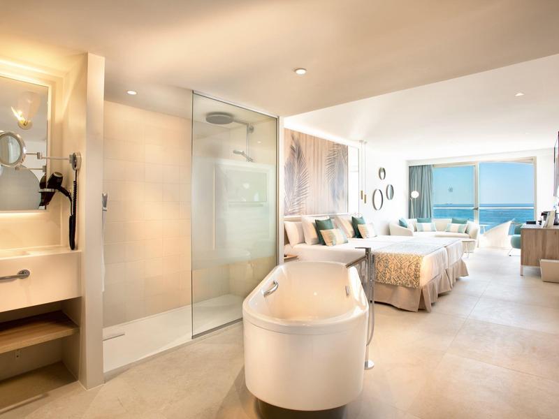 Helles Hotelzimmer mit Doppelbett, freistehender Wanne, Glasdusche und Meerblick durch große Fenster.