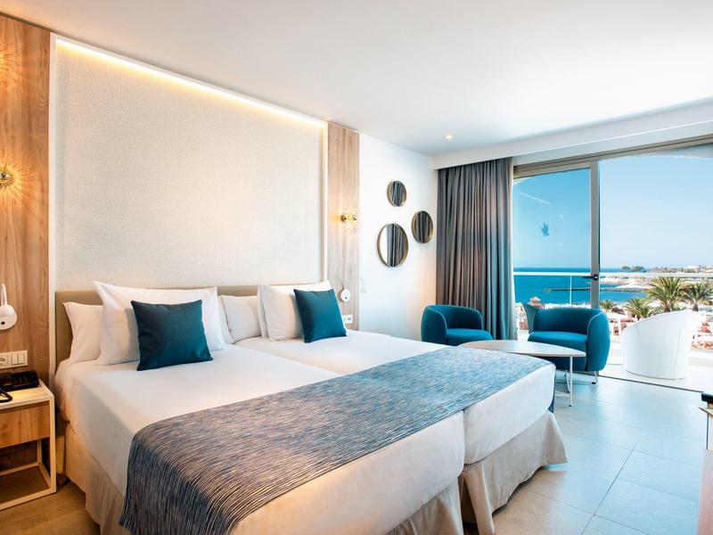 Helles, modernes Hotelzimmer mit zwei Betten, blauen Akzenten und großem Fenster zum Balkon mit Meerblick.