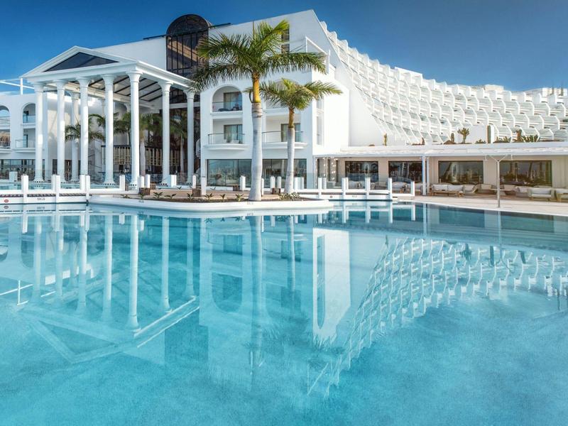 Großes weißes Hotel mit Palmen und klarem blauen Pool unter blauem Himmel.