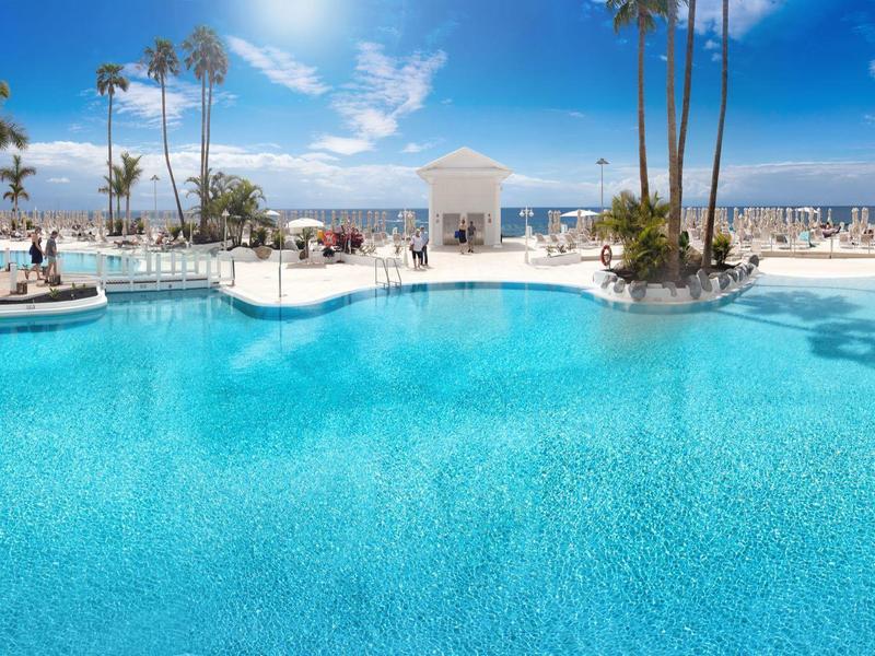Großer blauer Pool mit Palmen, weißen Liegen und klarem Himmel am sonnigen Strand.