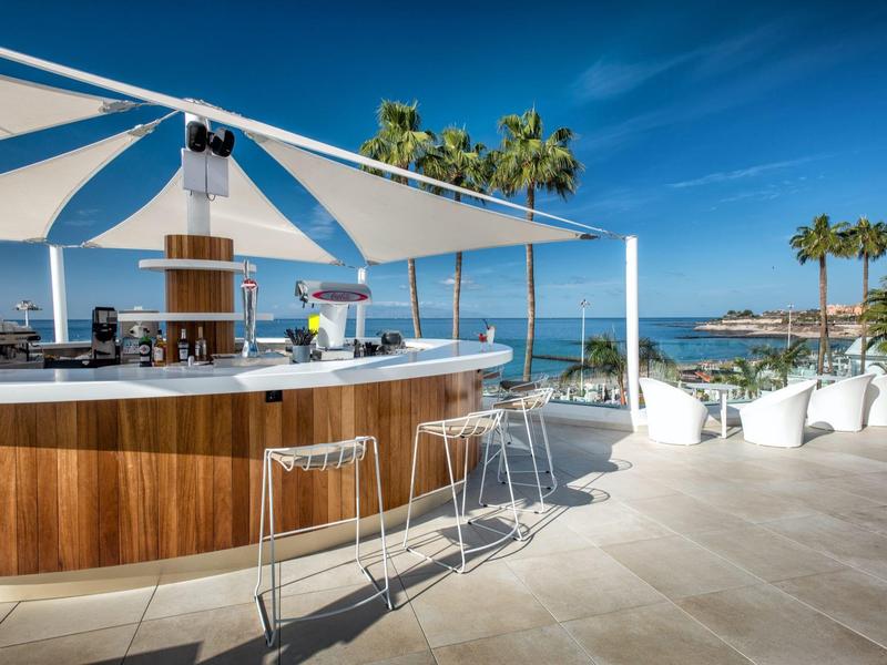 Moderner Außenbarbereich mit Holztheke, weißen Stühlen und Segel-Sonnenschutz am Meer bei blauem Himmel.