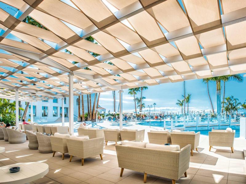 Terrasse mit weißen Loungemöbeln und Pergola, Blick auf Pool, Palmen und blaues Meer im Hintergrund.