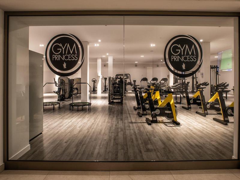 Moderner Fitnessraum mit Geräten und Holzfußboden, sichtbar durch große Glasfenster.