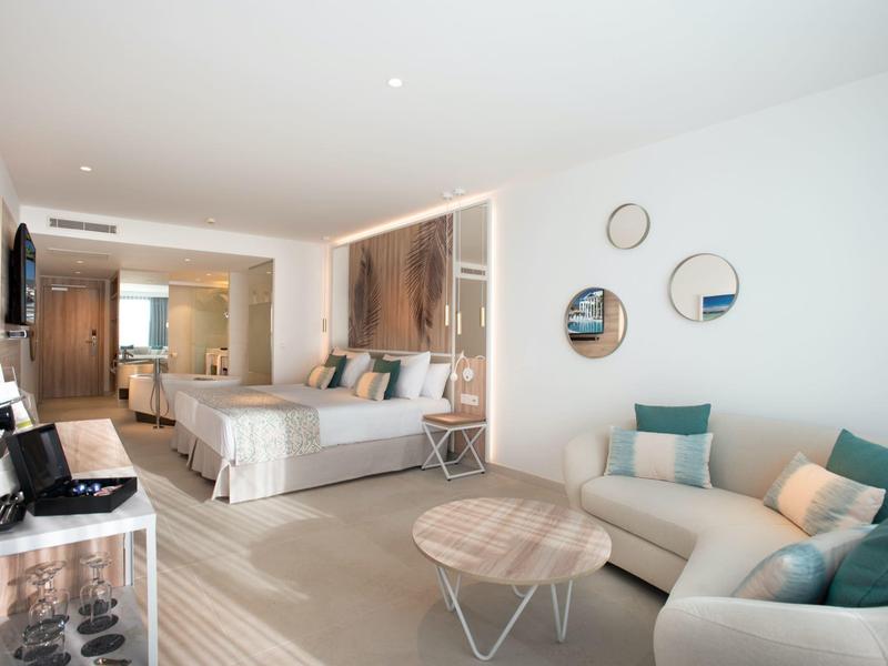 Helles Hotelzimmer mit großem Bett, Sofa, Tischen, Wandspiegeln und modernem Holzfußboden.