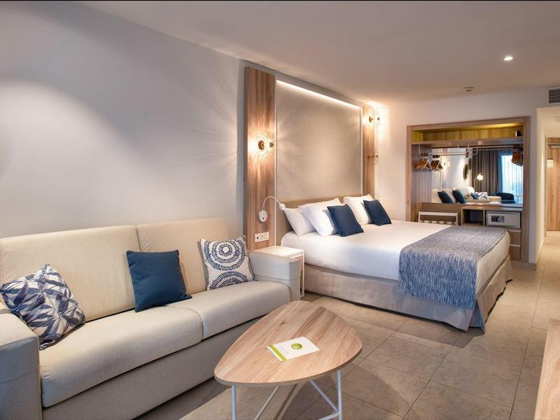 Modern eingerichtetes Hotelzimmer mit Doppelbett, Sofa, Holztisch und großer Spiegelwand.