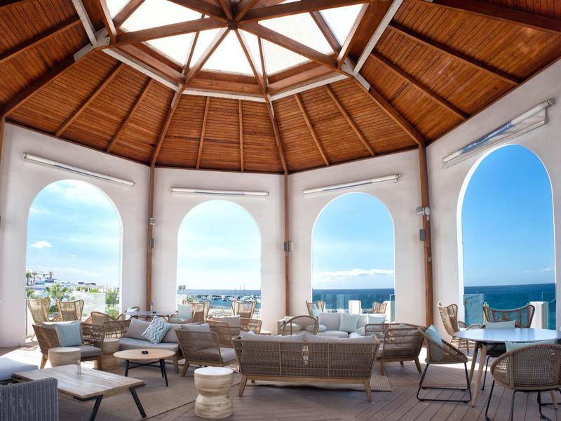 Offener Raum mit Holzdach, großen Bögen und Blick auf das Meer mit Sitzgruppen und Tischen.