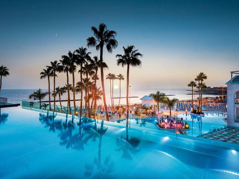 Großer Pool mit Palmen bei Sonnenuntergang, gemütliche Sitzbereiche und hell erleuchtetes Hotelgebäude.