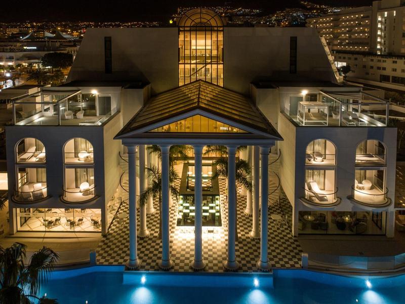 Großes weißes Gebäude mit geschwungenen Fenstern und beleuchtetem Pool bei Nacht.