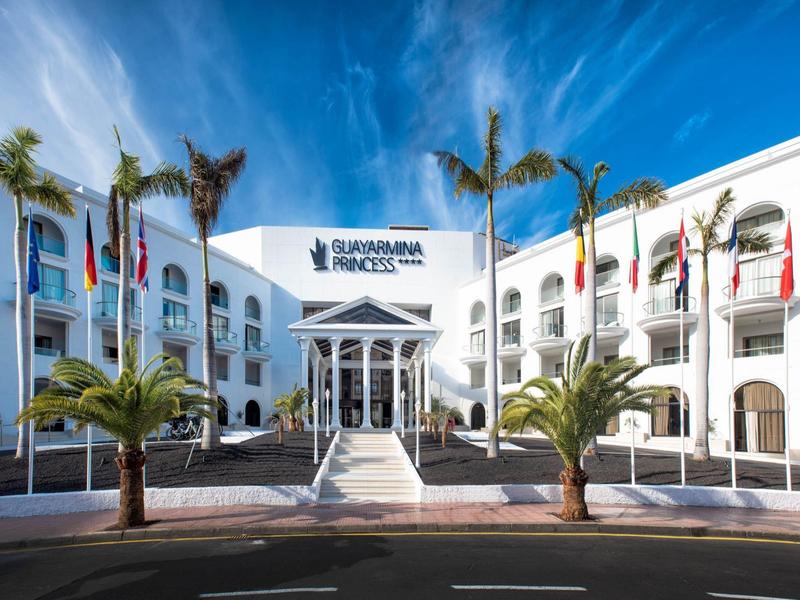 Großes weißes Hotelgebäude mit Palmen, vielen Flaggen und blauem Himmel im Hintergrund.