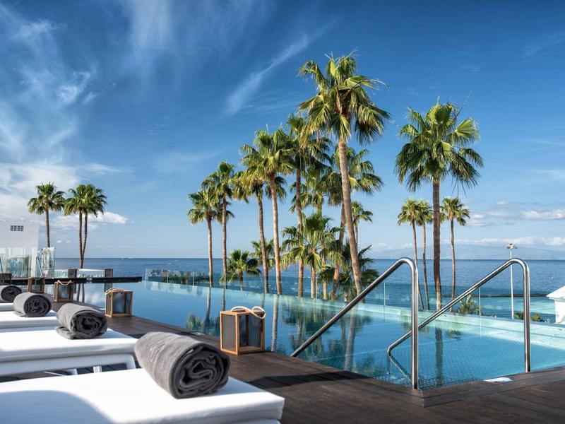 Luxuriöser Poolbereich mit Liegen, Palmen und Blick aufs blaue Meer und den Himmel.