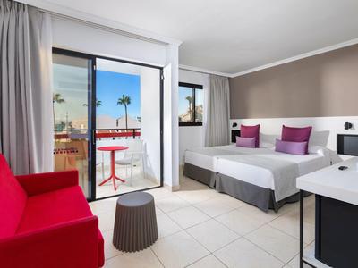 Camera d'hotel luminosa con letto matrimoniale, divano rosso e balcone con vista su palme.