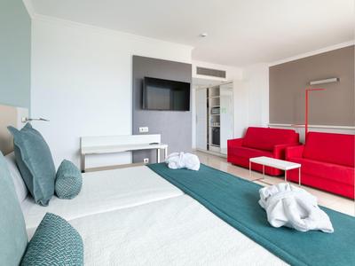 Camera d'hotel moderna con due letti singoli, divano rosso e TV a schermo piatto a muro.