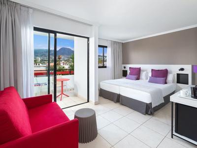 Camera d'hotel luminosa con letto matrimoniale, divano rosso e balcone con vista montagna.