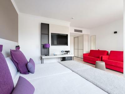 Camera d'hotel moderna con letto bianco, cuscini viola e divano rosso in ambiente luminoso.