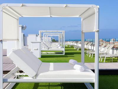 Lettini bianchi con baldacchino su prato sintetico verde accanto alla piscina sotto un cielo blu.