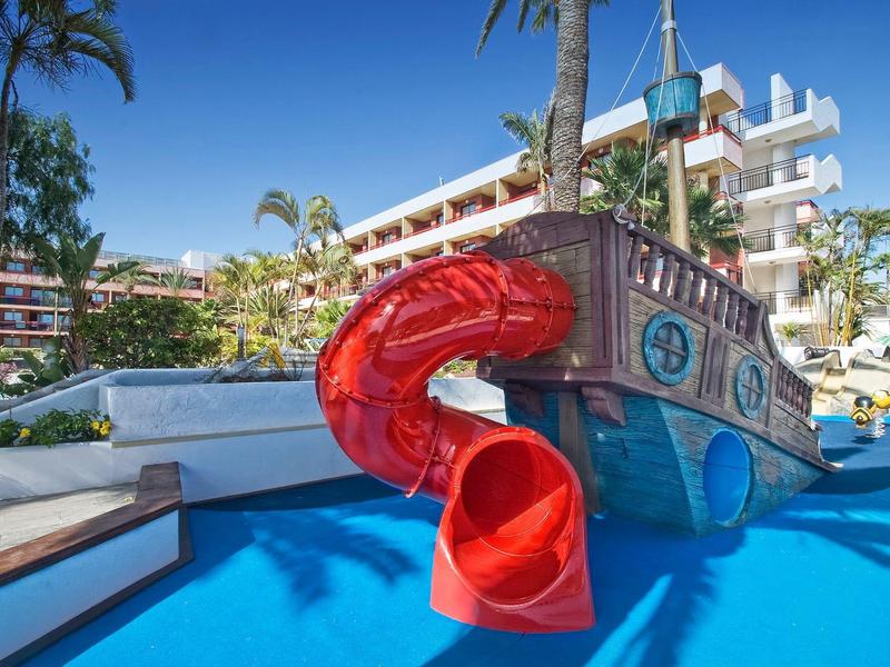 Colorata nave pirata con scivolo rosso nella piscina dell'hotel, circondata da palme.