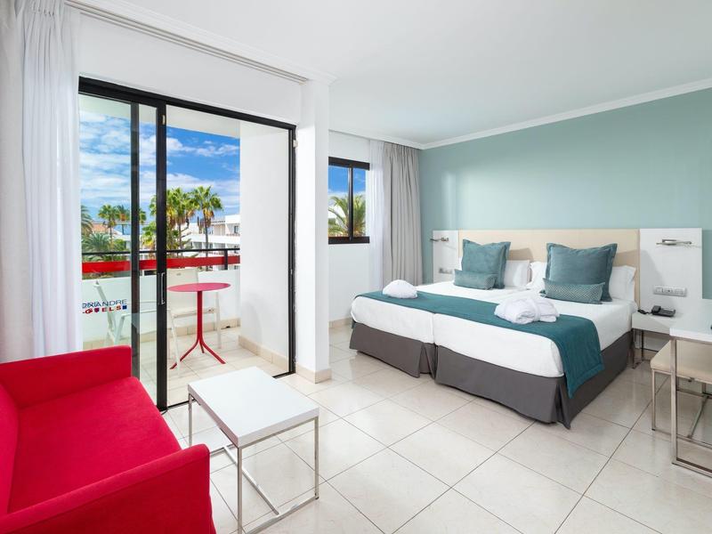 Camera d'hotel luminosa con letto matrimoniale, poltrona rossa e balcone con tavolo e sedia.