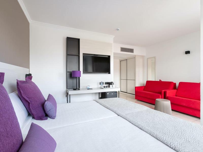 Camera d'hotel moderna con letto bianco, cuscini viola e divano rosso in ambiente luminoso.