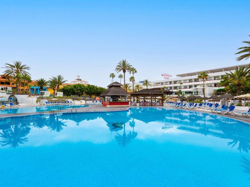 Grande piscina dell'hotel con acqua blu, ombrelloni e palme sotto un cielo limpido.