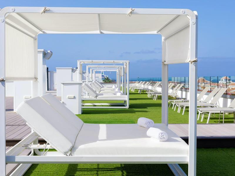 Lettini bianchi con baldacchino su prato sintetico verde accanto alla piscina sotto un cielo blu.