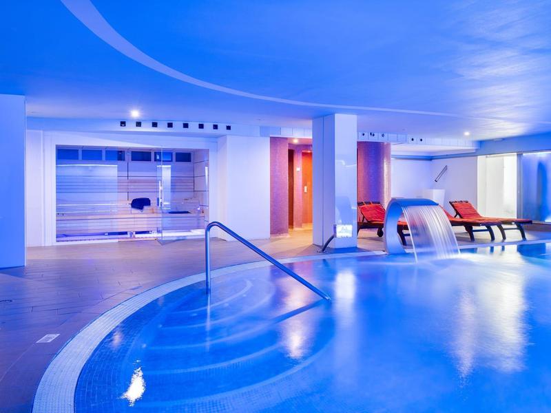 Vista interna di una piscina d'hotel moderna e illuminata con sedie a sdraio e getti d'acqua.