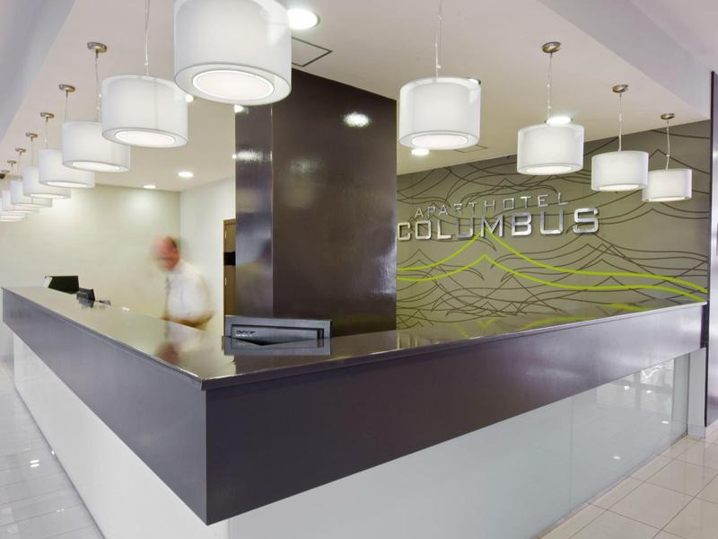 Moderner Empfangsbereich mit großem dunklem Tresen, weißen Lampen und grünem Hintergrund mit Schriftzug.