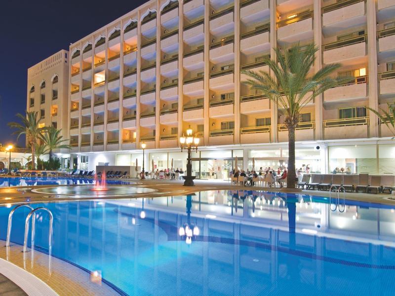 Großer beleuchteter Hotelpool mit blauer Wasseroberfläche und mehreren Palmen bei Nacht.