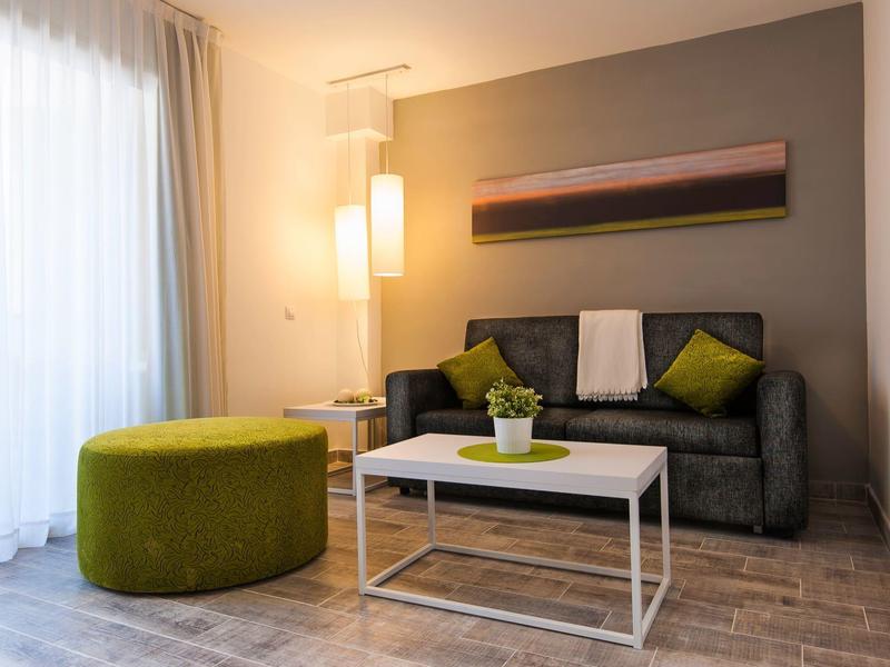 Schlichtes Wohnzimmer mit grauer Couch, grünem Pouf, weißem Tisch und großem Fenster mit Vorhängern.