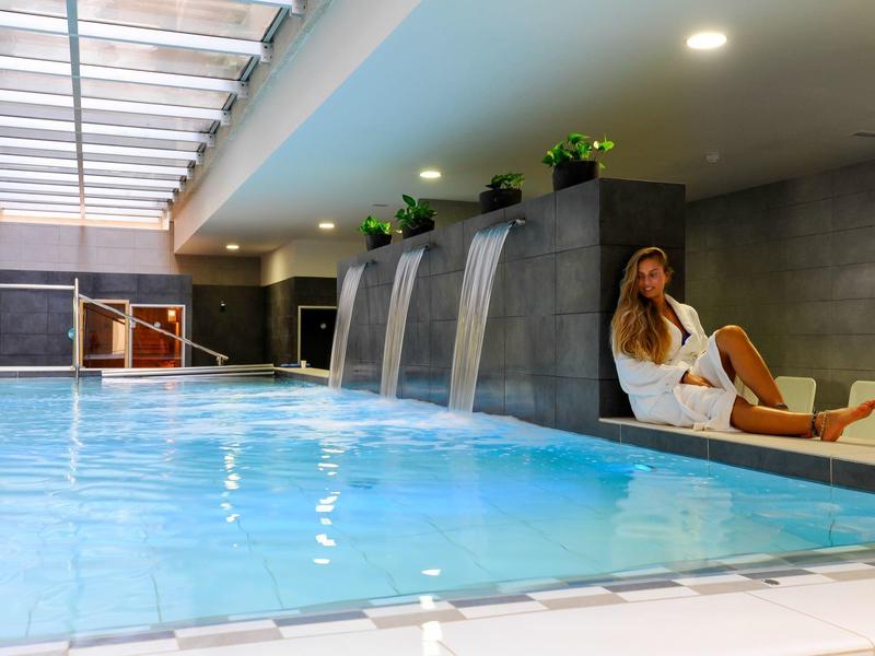 Moderner Innenpool mit Wasserfällen, blauen Fliesen und einer Frau in Bademantel auf Liege.