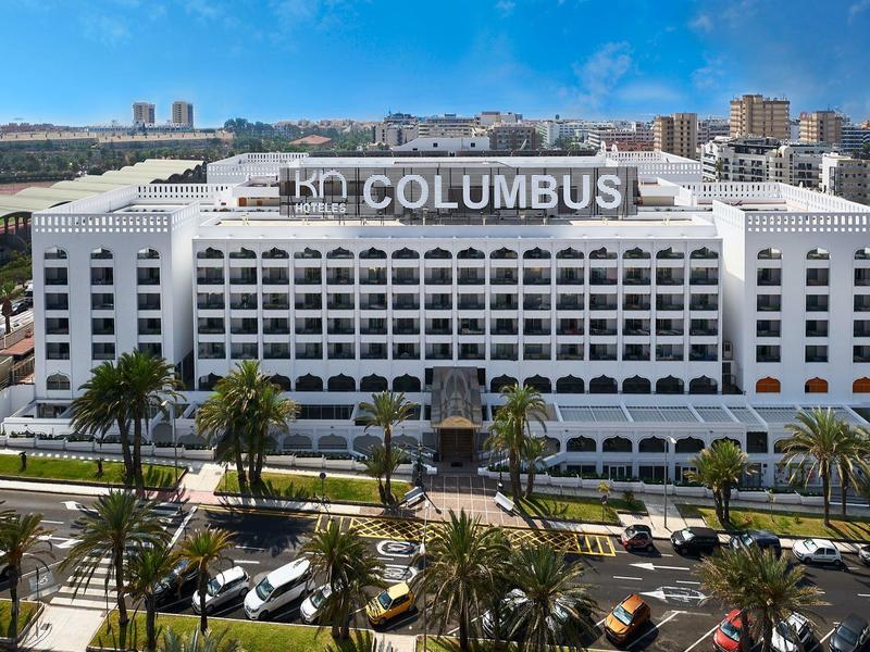 Großes weißes Hotelgebäude mit vielen Balkonen vor blauem Himmel und Palmen in der Stadt.