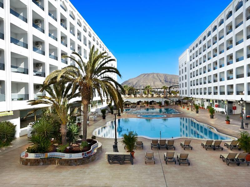 Großes Hotel mit Swimmingpool zwischen zwei Gebäuden, Palmen und Liegestühlen bei klarem Himmel.