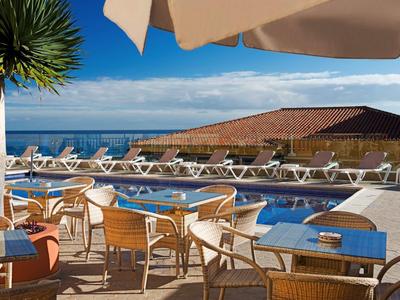Außenbereich mit Pool, Holztischen, Stühlen und Sonnenschirmen, Blick auf Meer und blauen Himmel.