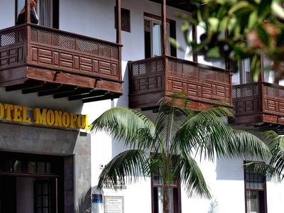 Weißes Hotelgebäude mit braunen Balkonen und Palmen davor, Sonnenschild gelb mit schwarzer Schrift.
