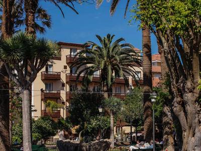 Edificio de hotel con balcones, rodeado de palmeras y jardín cuidado bajo cielo azul